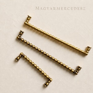 Può includere: Tre maniglie per cassetti in metallo color ottone di diverse lunghezze, ciascuna caratterizzata da un design a perline sferiche ripetute lungo l'impugnatura e piccole piastre di montaggio circolari alle estremità. Il testo MAGYARMERCEDESZ è in alto.