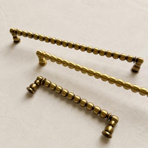 May include: Three brass cabinet pulls of varying lengths, each featuring a continuous row of spherical beads along the handle and rounded mounting posts at both ends. The metal has a matte, brushed finish with a warm, golden-brown metallic tone.
