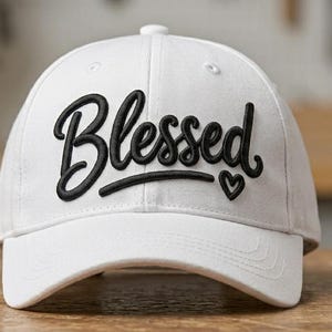 May include: A white baseball cap featuring the word Blessed embroidered in black script font across the front panels. The embroidery includes a small black heart symbol positioned to the right of the text. The cap has a structured crown and a curved brim.