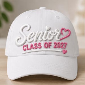 May include: A white baseball cap featuring embroidered text that reads Senior in white script with a pink heart, and CLASS OF 2027 in pink block letters below it. A small pink heart is positioned at the end of a horizontal line under the text.