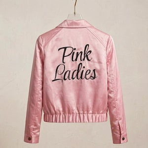 Può includere: Un giubbotto bomber in raso rosa chiaro con colletto, maniche lunghe con polsini abbottonati e vita elasticizzata. Sul retro è presente la scritta Pink Ladies ricamata in corsivo nero, esposta su una gruccia contro una parete neutra.
