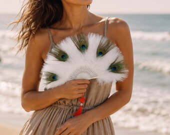 May include: A white hand fan with peacock feathers and a red tassel. The fan has a semi-circular shape and is held in front of a person wearing a tan dress. The background is a beach with the ocean.