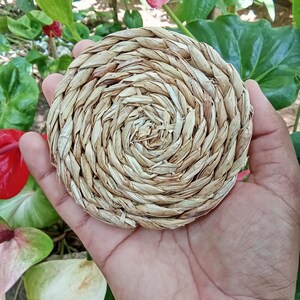 May include: A circular coaster crafted from braided natural dried straw or seagrass fibers. The coaster features a tight, concentric spiral pattern with a light tan and beige color palette, showcasing a textured, woven construction throughout.