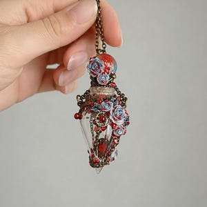 Può includere: Una collana con pendente in vetro realizzata a mano con una fiala trasparente a forma di goccia. La fiala è adornata con rose rosse e blu, gemme rosse e dettagli in metallo color bronzo. Il pendente è appeso a una catena di bronzo.