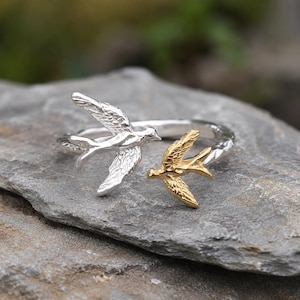 May include: An open-style ring featuring two bird motifs. One bird is finished in a polished silver tone, while the smaller bird is finished in a gold tone. Both birds have textured wing details and are positioned to face each other on the band.