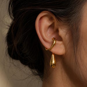 Puede incluir: Un pendiente tipo ear cuff de tono dorado con un acabado liso y pulido que rodea el cartílago. Un dije sólido en forma de gota, también de tono dorado, cuelga de la base del pendiente, creando una apariencia metálica y reflectante.