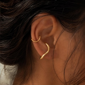 May include: Two gold-toned ear cuffs worn on the cartilage and conch of an ear. The upper cuff is a simple, smooth, thin circular band. The lower cuff features a thicker, wavy, organic silhouette that contours along the outer edge of the ear.
