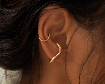 May include: Two gold-toned ear cuffs worn on the cartilage and conch of an ear. The upper cuff is a simple, smooth, thin circular band. The lower cuff features a thicker, wavy, organic silhouette that contours along the outer edge of the ear.