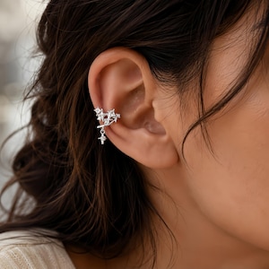 May include: A silver-toned ear cuff featuring a cluster of small, clear, square-cut stones arranged in a star-like formation. A single, dangling, four-pointed star charm set with a clear stone hangs from the main body of the ear cuff.