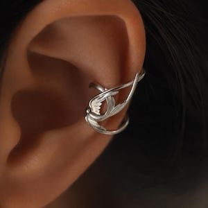 May include: A silver ear cuff featuring a detailed bird design with outstretched wings. The metal piece wraps around the cartilage of the ear, showcasing intricate feather textures and a polished finish that catches the light along its curved form.