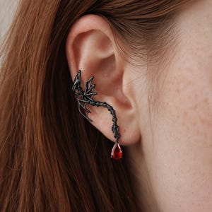 Può includere: Un ear cuff in metallo nero a forma di drago con ali spiegate e una lunga coda segmentata. Il drago presenta piccoli accenti rossi sulle ali e una singola gemma rossa a forma di goccia sospesa all'estremità della coda.