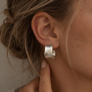 Könnte beinhalten: Ein silberfarbener Creolen-Ohrring mit einem breiten, geschwungenen und leicht unregelmäßigen Banddesign. Die Oberfläche des Ohrrings hat ein mattes, gebürstetes Finish, das ein weiches metallisches Aussehen erzeugt und am Ohrläppchen anliegt.