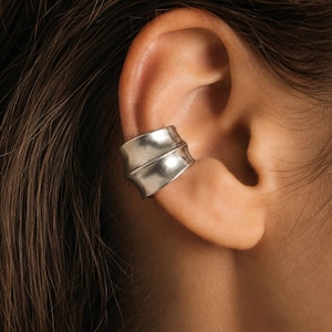 May include: A wide, silver-toned ear cuff featuring a smooth, curved surface with a central horizontal ridge. The metal has a matte, slightly weathered finish and wraps around the outer edge of the ear cartilage, creating a layered appearance.