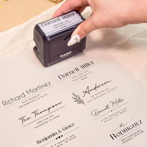 May include: A black Trodat 4913 self-inking rubber stamp being pressed onto paper. The stamp features the name Darnell Miller and a California address. The paper displays multiple printed address labels with various names and locations in black ink.