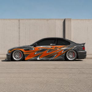May include: A grey sports car featuring orange and white flame decals along its side. The car has custom rims with red brake calipers. The vehicle is parked on a paved surface in front of a concrete wall.
