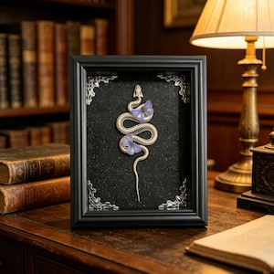 May include: A black rectangular shadow box frame containing a white snake skeleton sculpture adorned with two light purple butterflies. The frame features ornate silver corner accents and a dark, speckled background, creating a high-contrast display.