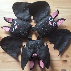 May include: Three handmade felt bat plushies arranged in a circular pattern. Each bat is black with pink inner ears, white stitched details, and white felt eyes. Two bats have open wings, while the third bat is positioned with its wings folded.