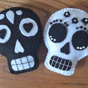 May include: Two felt skull decorations, one black with white accents and heart-shaped eye sockets, the other white with black eyes and floral embellishments. Both have stitched details and toothy grins.