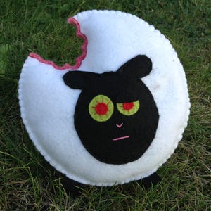 Può includere: Un peluche rotondo in feltro bianco a forma di pecora con un grande segno di morso sul bordo superiore. La pecora ha un viso nero, due piccoli piedi neri e due occhi con iridi verde lime e pupille rosse.