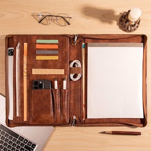 May include: Open brown leather portfolio with a zipper closure, containing a notepad, phone, pens, and cards. A laptop, glasses, and a candle holder are on the wooden surface.