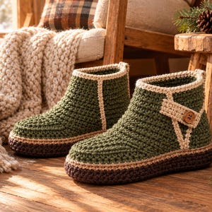 May include: A pair of ankle-high crochet slippers featuring a dark olive green upper, a beige trim along the seams, and a dark brown sole. Each slipper includes a decorative beige strap across the side secured with a small brown wooden button.