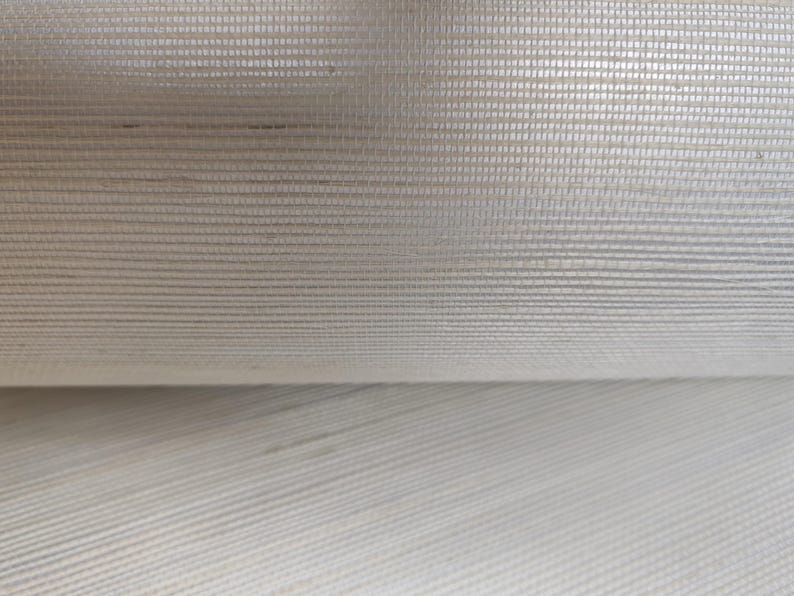 Op de afbeelding: Een rol gebroken wit behang met een fijne horizontale en verticale rasterstructuur. Het materiaal heeft een subtiele glans en een geweven uiterlijk, waarbij zichtbare vezels een consistent, herhalend patroon over het oppervlak cre&euml;ren.