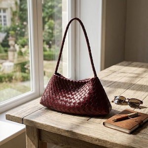 May include: A burgundy leather handbag with a woven design sits on a wooden table. The bag has a braided handle and a triangular shape. Sunglasses and a leather-bound notebook with a pen are also on the table.