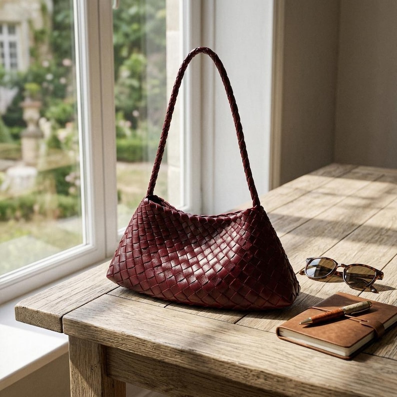 May include: A burgundy leather handbag with a woven design sits on a wooden table. The bag has a braided handle and a triangular shape. Sunglasses and a leather-bound notebook with a pen are also on the table.
