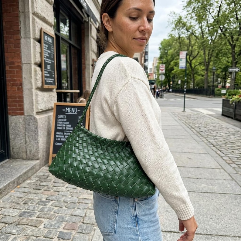 May include: A green woven leather shoulder bag with a braided strap. The bag is rectangular and has a textured appearance. The woman is wearing a cream-colored sweater and blue jeans.