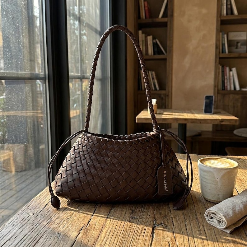 May include: A brown woven leather handbag with a braided handle. The bag has a rectangular shape with a rounded bottom and a small tag that says "MENG LI SHA". The bag is sitting on a wooden table.