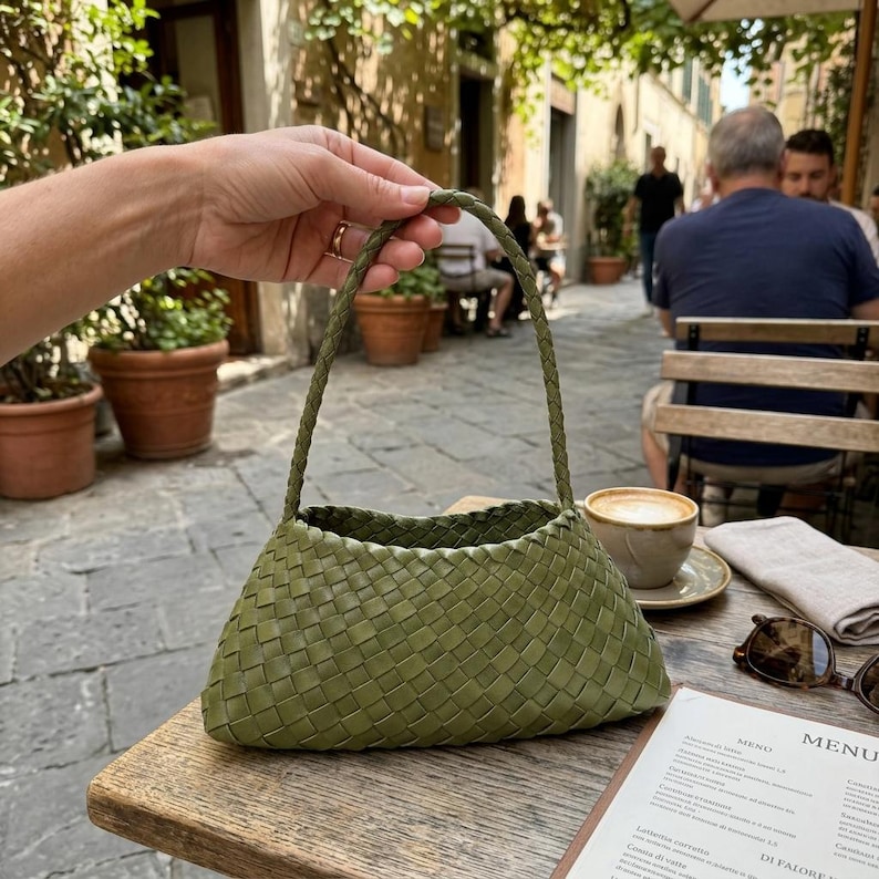 May include: A woven olive green handbag with a braided handle. The bag has a trapezoid shape and is held by a person. The background shows a cafe setting with a coffee cup, menu, and sunglasses on a wooden table.