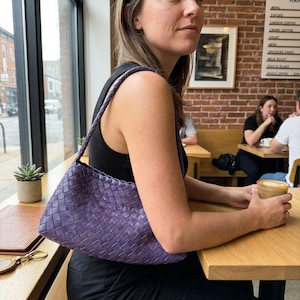 May include: A woven, purple shoulder bag with a braided strap. The bag is held by a person sitting at a wooden table in a cafe. The bag's texture is visible, and the color is a deep, saturated purple.