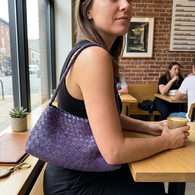 May include: A woven, purple shoulder bag with a braided strap. The bag is held by a person sitting at a wooden table in a cafe. The bag's texture is visible, and the color is a deep, saturated purple.