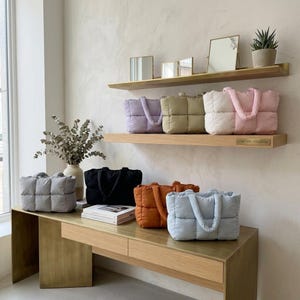 Peut inclure: Présentation de sacs fourre-tout matelassés de différentes couleurs : lavande, vert olive, crème, rose, gris, noir, orange et bleu clair. Les sacs sont disposés sur des étagères et une table en bois, mettant en valeur leur design et leurs couleurs.