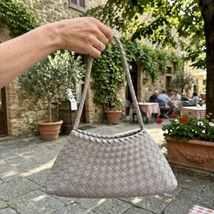 May include: A light gray woven leather handbag with braided handles. The bag has a trapezoid shape and is held up by a person's hand. The background shows a stone building and outdoor seating.