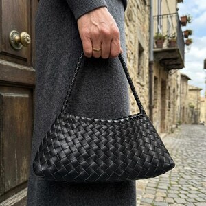 May include: A black woven leather handbag with a braided handle. The bag has a rectangular shape with a curved top and is held by a person. The background shows a cobblestone street and buildings.