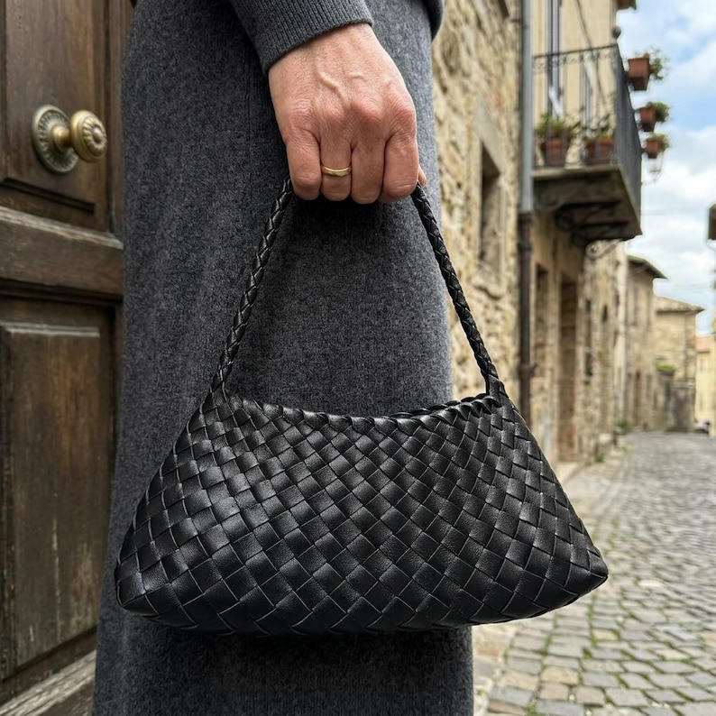 May include: A black woven leather handbag with a braided handle. The bag has a rectangular shape with a curved top and is held by a person. The background shows a cobblestone street and buildings.