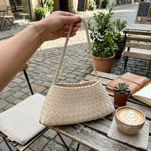 May include: A beige woven handbag with a braided handle is held up in front of a cafe table. The bag has a triangular shape. A cup of coffee with latte art, a small succulent, and a notebook are on the table.