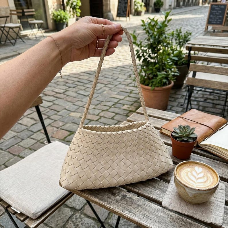 May include: A beige woven handbag with a braided handle is held up in front of a cafe table. The bag has a triangular shape. A cup of coffee with latte art, a small succulent, and a notebook are on the table.