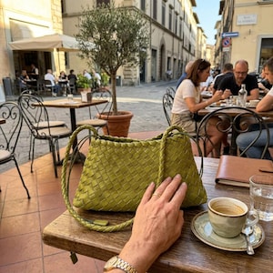 May include: A woven, olive-green handbag with braided handles rests on a wooden table. A cup of coffee sits on a saucer nearby. The scene is set outdoors at a cafe, with people seated at tables in the background.