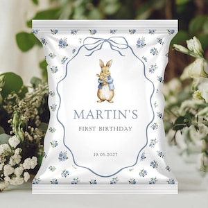 Peut inclure: Un sachet de fête blanc avec un motif floral bleu et un motif de ruban bleu. Le sachet présente une illustration à l'aquarelle d'un lapin tenant un animal en peluche. Le texte indique "MARTIN'S FIRST BIRTHDAY" et la date "19.05.2027".