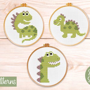 May include: Three mini cross-stitch patterns of green dinosaurs in wooden hoops. The dinosaurs have purple accents and are displayed on a light wood background. The text "mini patterns" and "23 x 34 Stitches" are also visible.
