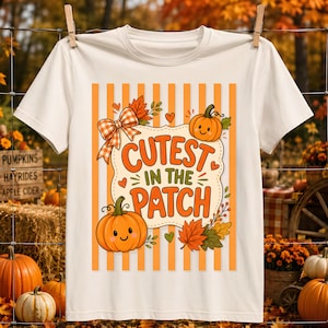 May include: A cream-colored short-sleeve t-shirt featuring a graphic print of orange vertical stripes, two smiling pumpkins, autumn leaves, and a plaid bow. The central text reads "CUTEST IN THE PATCH" in bold orange letters on a scalloped background.