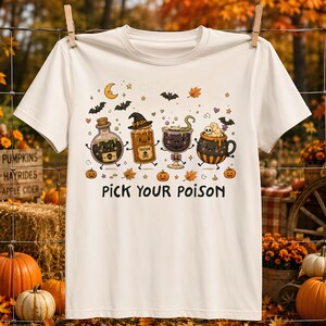 May include: A cream-colored short-sleeve t-shirt featuring a graphic print of four animated Halloween-themed containers, including a potion bottle, a jar, a goblet, and a cauldron, with the text PICK YOUR POISON printed in black below them.