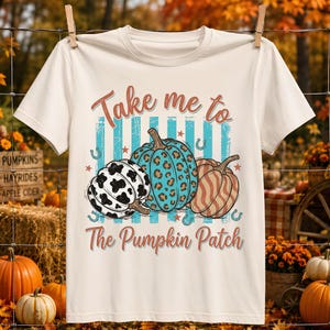 Könnte beinhalten: Ein cremefarbenes Kurzarm-T-Shirt mit einer Grafik aus drei Kürbissen mit Kuhflecken-, Leoparden- und Streifenmuster. Der Text Take me to The Pumpkin Patch ist in rostfarbener Schrift gehalten und mit kleinen Sternen und Hufeisen verziert.