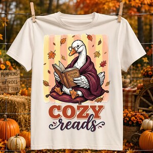 Puede incluir: Una camiseta color crema con el gráfico de un ganso blanco que lleva gafas y una manta morada oscura mientras lee un libro titulado Autumn Tales. El diseño incluye hojas de otoño y el texto Cozy reads en colores naranja y morado.