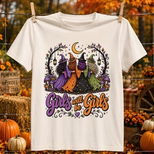 Könnte beinhalten: Ein cremefarbenes Kurzarm-T-Shirt mit einer Grafik von vier Hexen in bunten Gewändern unter einem Halbmond. Der Text "Girls will be Girls" ist unter der Illustration in violetter und orangefarbener Schreibschrift aufgedruckt.