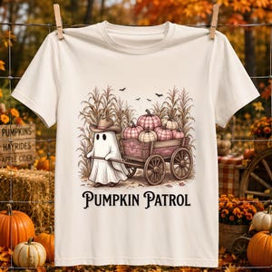 Può includere: Una t-shirt color crema con l'illustrazione di un fantasma con cappello da cowboy che traina un carro di legno pieno di zucche a quadri rosa e bianchi in un campo di mais. Il testo PUMPKIN PATROL è stampato in nero sotto l'illustrazione.