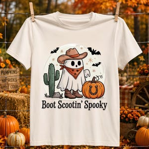 Könnte beinhalten: Ein cremefarbenes Kurzarm-T-Shirt mit einer Grafik eines Geistes, der einen braunen Cowboyhut, ein gemustertes Halstuch und braune Stiefel trägt. Der Geist steht neben einem grünen Kaktus und einem orangefarbenen Kürbis mit Hufeisenmotiv. Text: Boot Scootin' Spooky.