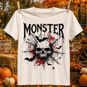 May include: A cream-colored short-sleeve t-shirt featuring a black graphic print of a skull centered within a spider web. The design includes flying bats, a small spider, and red paint splatters. The word MONSTER is printed in a bold, dripping font above the skull.